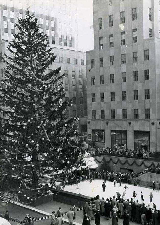 Black-and-white photo of the Rockefeller Center Christmas Tree in 1955, decorated with lights and ornaments as crowds gather around the rink below.