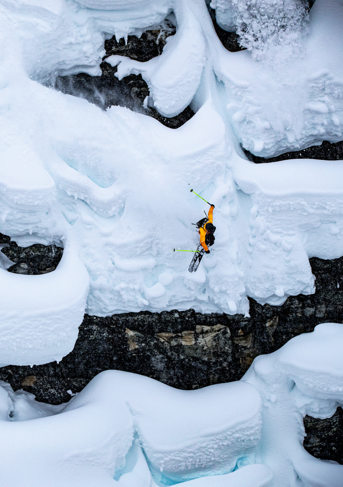 A high-angle, vertical photograph of a skier wearing a bright orange jacket performing a large drop or jump from a steep, rocky cliff face heavily covered in thick, sculpted white and blue-tinted snow. The skier is airborne, mid-tuck, with skis pointed down and poles held close, surrounded by overhanging snow formations and dark rock below.