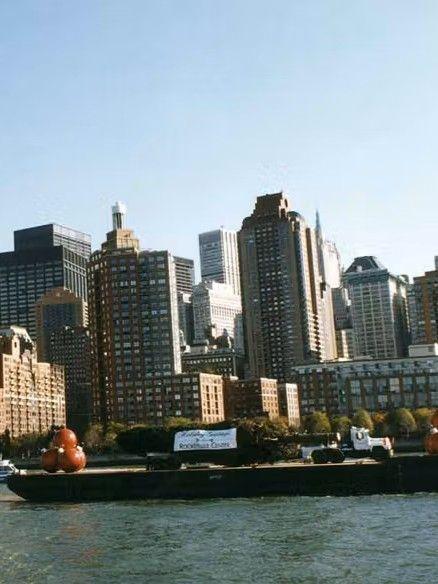 Color photo of a barge carrying the Rockefeller Center Christmas Tree along the Hudson River with the Manhattan skyline in the background.