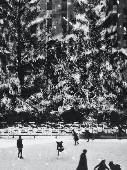 Black-and-white photo of skaters on the Rockefeller Center rink in 1936, with two Christmas trees and falling snow in the background.