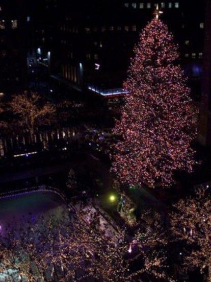 Nighttime photo of the Rockefeller Center Christmas Tree towering above the rink in 1999, illuminated with lights and surrounded by crowds.