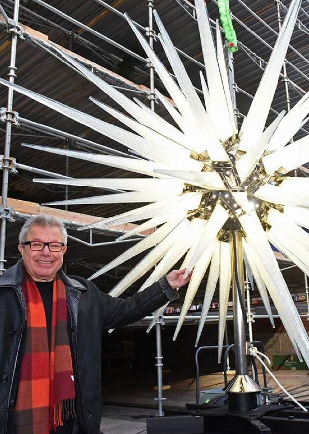 Color photo of architect Daniel Libeskind standing beside the redesigned Swarovski star, showing its illuminated crystal spikes before installation.