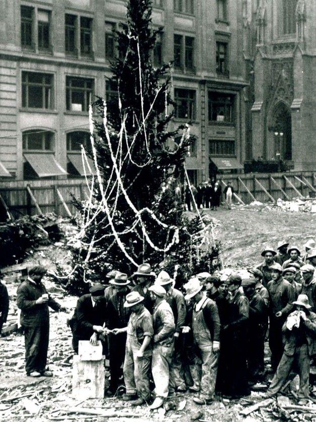 Black-and-white photo of construction workers gathered around the first Rockefeller Center Christmas tree in 1931, decorated with simple garlands.