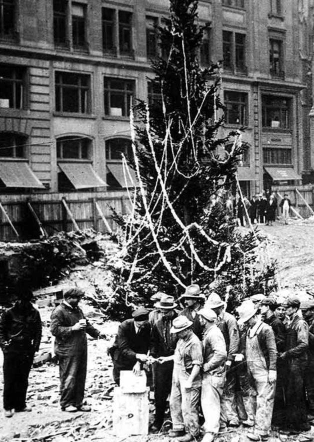 Black-and-white photo of construction workers gathered around the first Rockefeller Center Christmas tree in 1931, decorated with simple garlands.